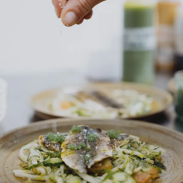 Une main saupoudre de sel un poisson grillé dressé sur un lit de légumes, second plat flou en arrière-plan.
