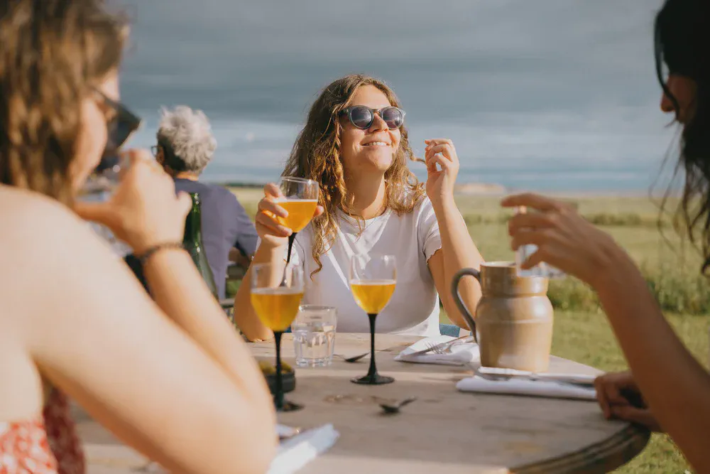 Un groupe partage un repas en extérieur, une femme en lunettes de soleil sourit un verre à la main, ciel nuageux.