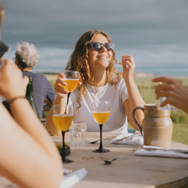 Un groupe partage un repas en extérieur, une femme en lunettes de soleil sourit un verre à la main, ciel nuageux.