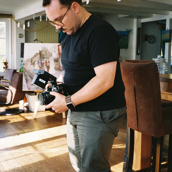 Un homme en tee-shirt noir examine un appareil photo à écran orientable, dans un salon décoré d’un tableau et de photos suspendues.
