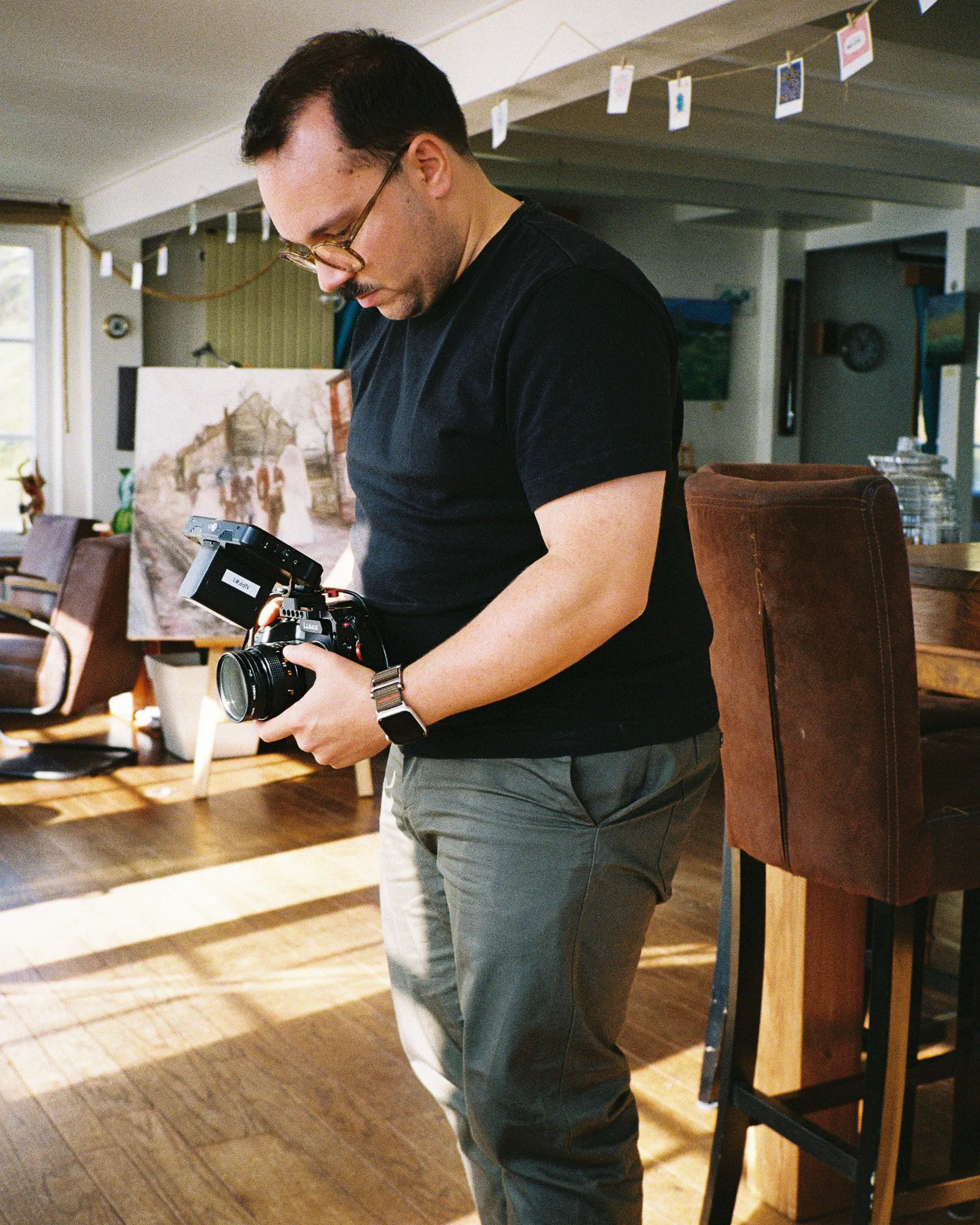 Un homme en tee-shirt noir examine un appareil photo à écran orientable, dans un salon décoré d’un tableau et de photos suspendues.