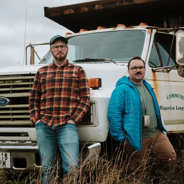 Deux hommes posent devant un vieux camion-benne rouillé marqué Commune de Miquelon-Langlade, ciel nuageux.