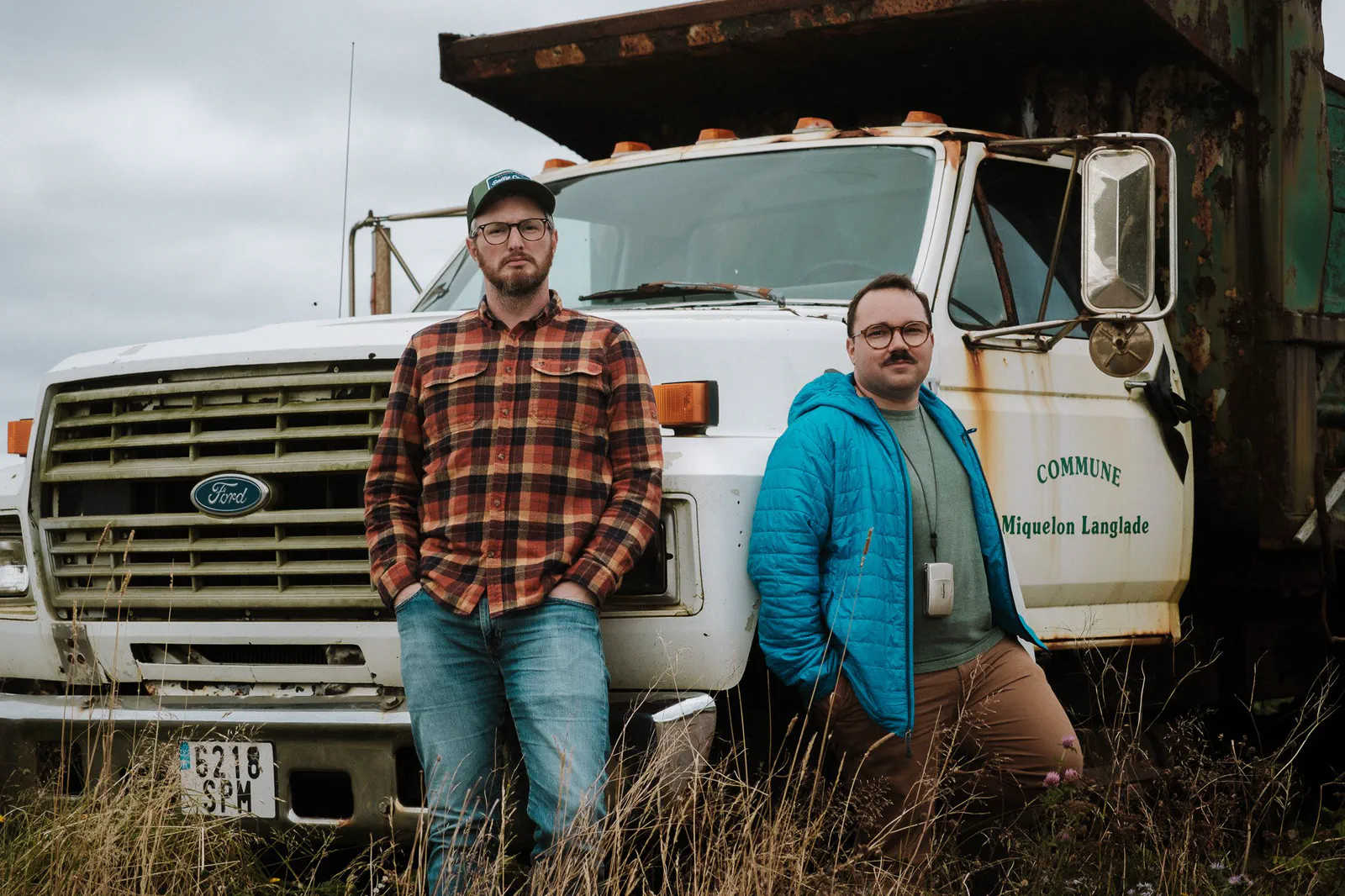 Deux hommes posent devant un vieux camion-benne rouillé marqué Commune de Miquelon-Langlade, ciel nuageux.
