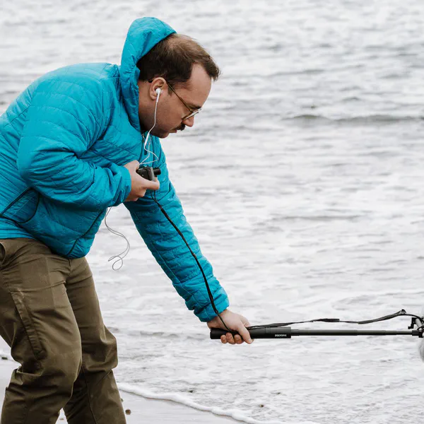 Un homme accroupi sur une plage tend un micro à bonnette vers les vagues, écouteurs aux oreilles.