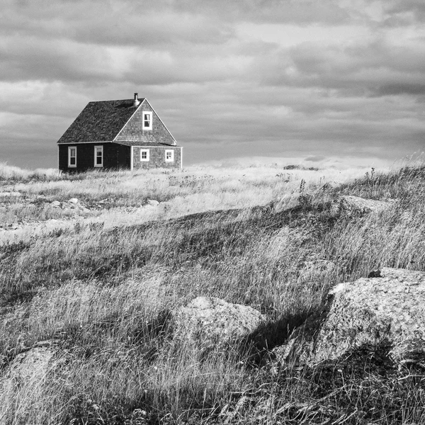Une petite maison sombre se dresse seule dans un paysage de dunes venteuses, rochers au premier plan, noir et blanc.