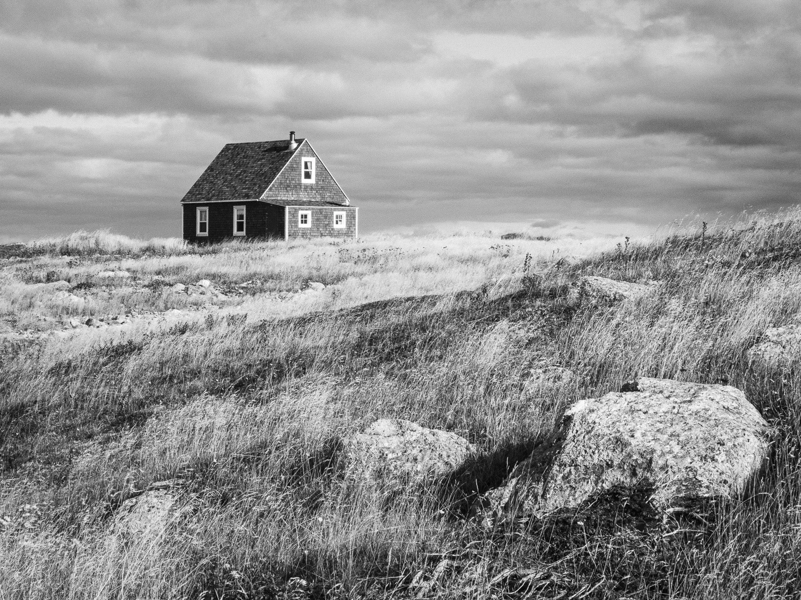 Une petite maison sombre se dresse seule dans un paysage de dunes venteuses, rochers au premier plan, noir et blanc.