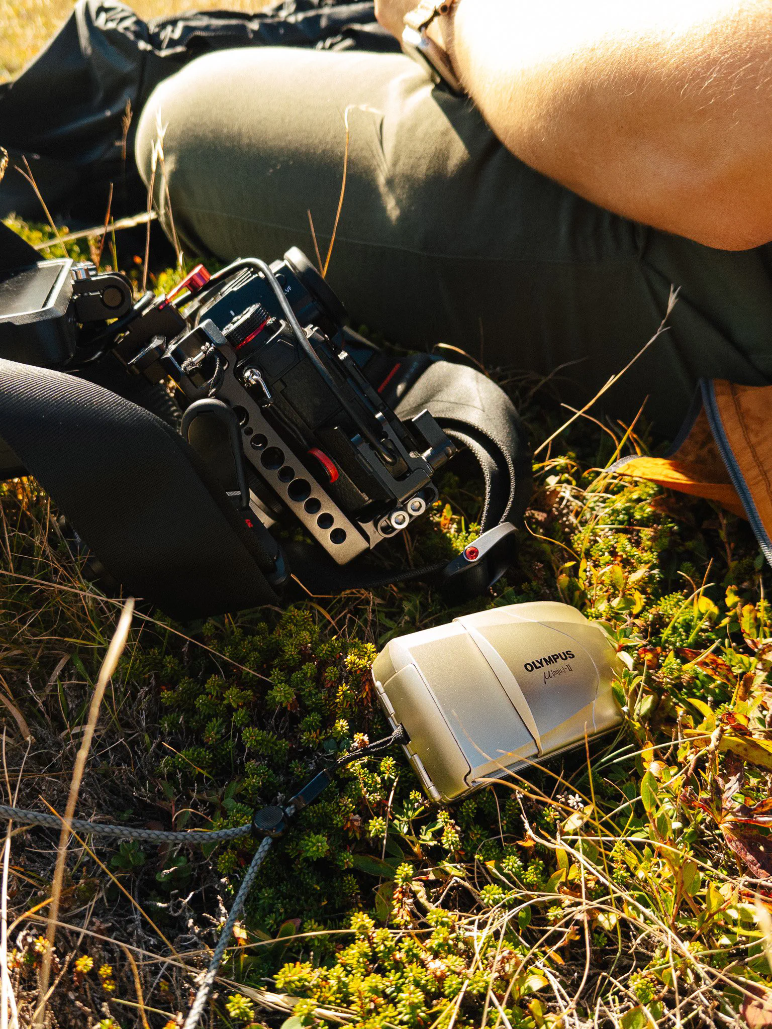 Gros plan sur un rig de caméra fixé à une jambe et un appareil photo compact argentique posé dans l’herbe.