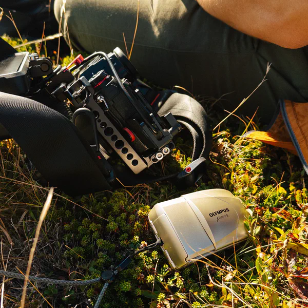 Gros plan sur un rig de caméra fixé à une jambe et un appareil photo compact argentique posé dans l’herbe.