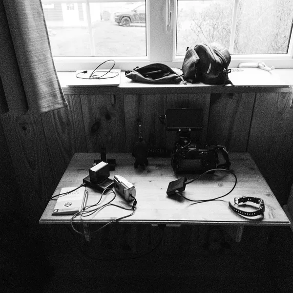 Du matériel vidéo est disposé sur une petite table sous une fenêtre, sacs posés sur le rebord, noir et blanc.