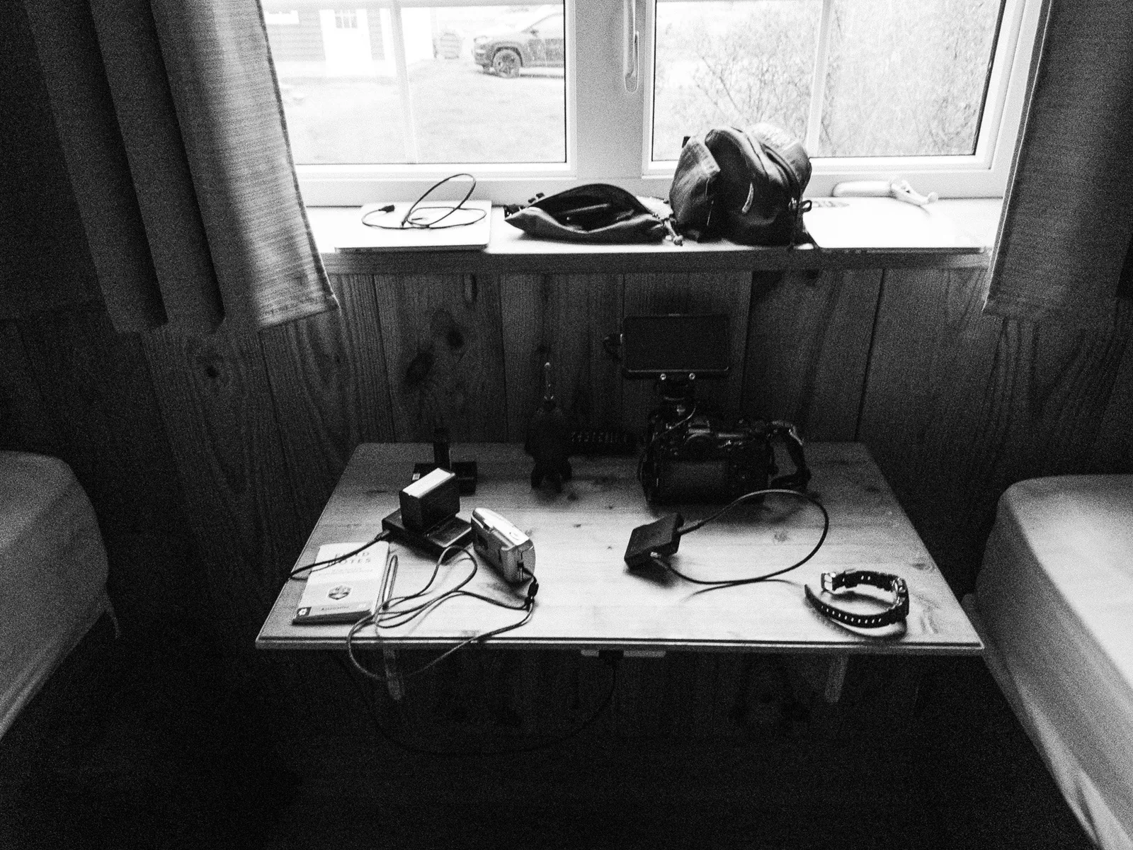 Du matériel vidéo est disposé sur une petite table sous une fenêtre, sacs posés sur le rebord, noir et blanc.